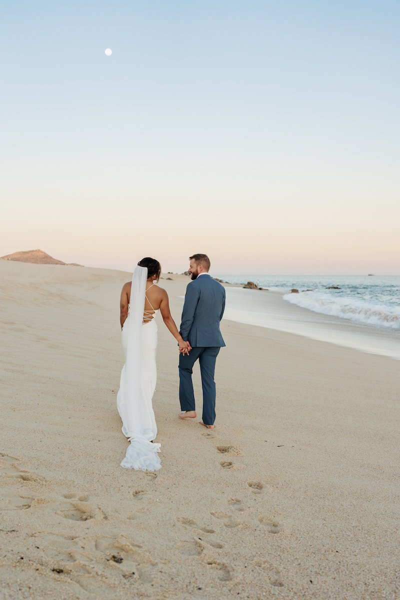 Wedding couple walking away from camera on beach, holding hands