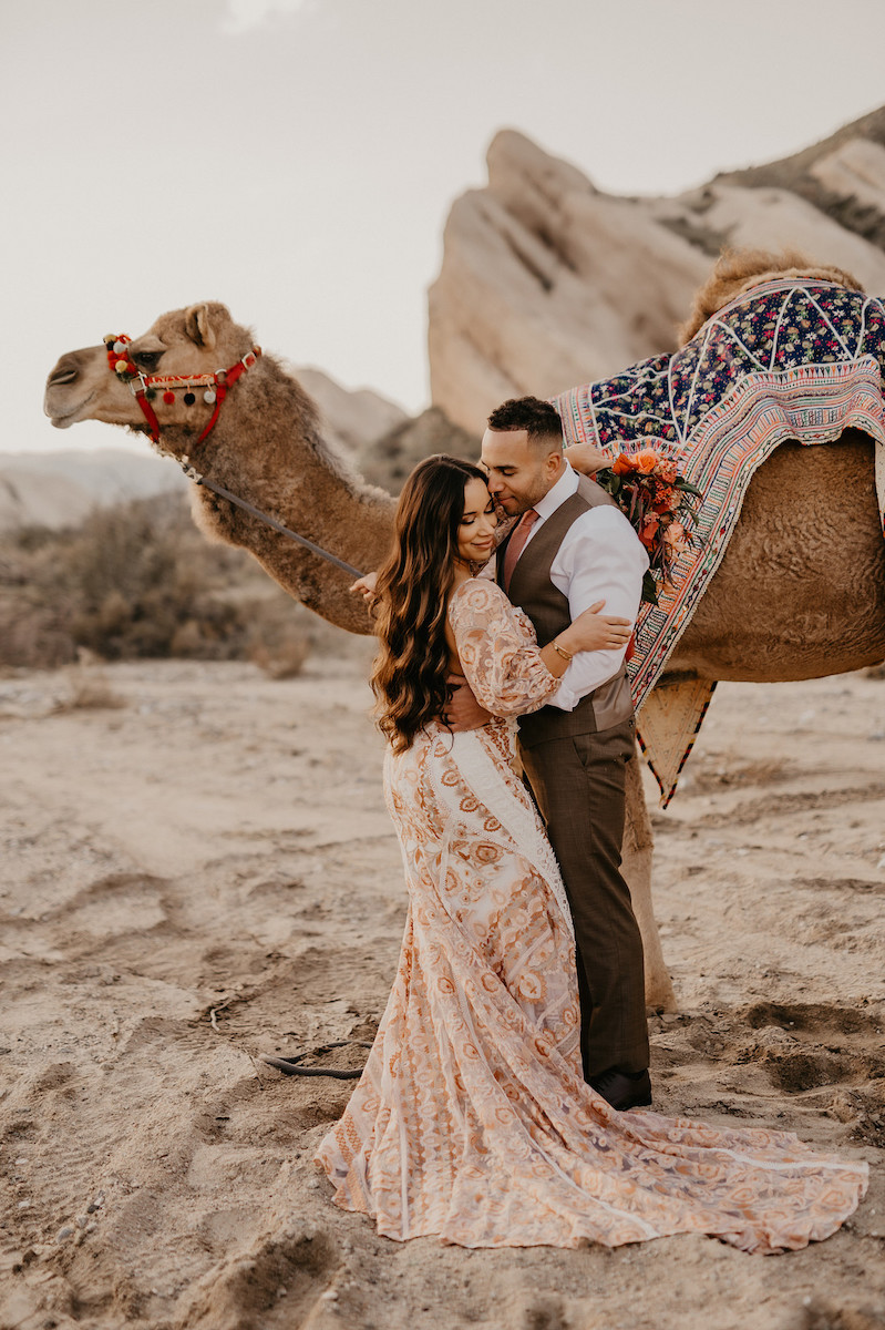 A bride in a lace dress with a groom kissing her in front of a camel in the desert