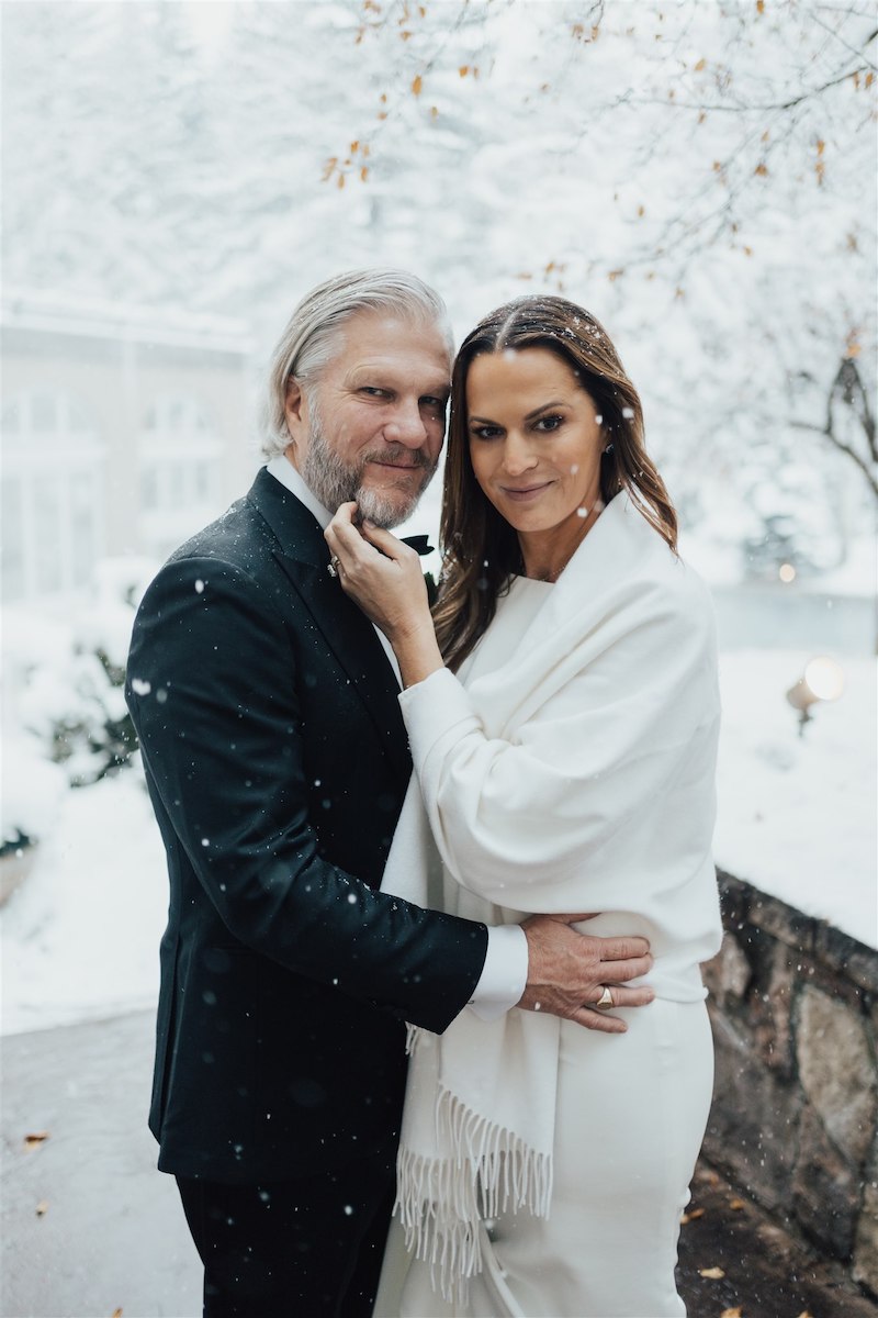 A couple dressed in formal attire poses affectionately in a snowy setting.
