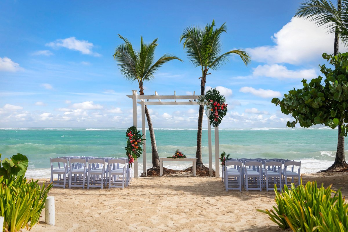 wedding ceremony set up on a beach with white folding chairs facing a wooden arch decorated with white flowers and greenery and turquoise ocean water