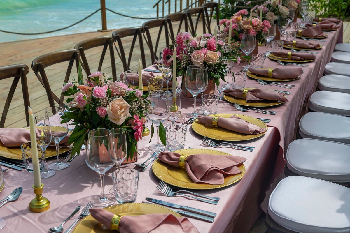 A long dinner table with pink florals and linens and gold accents set for a party on the deck.