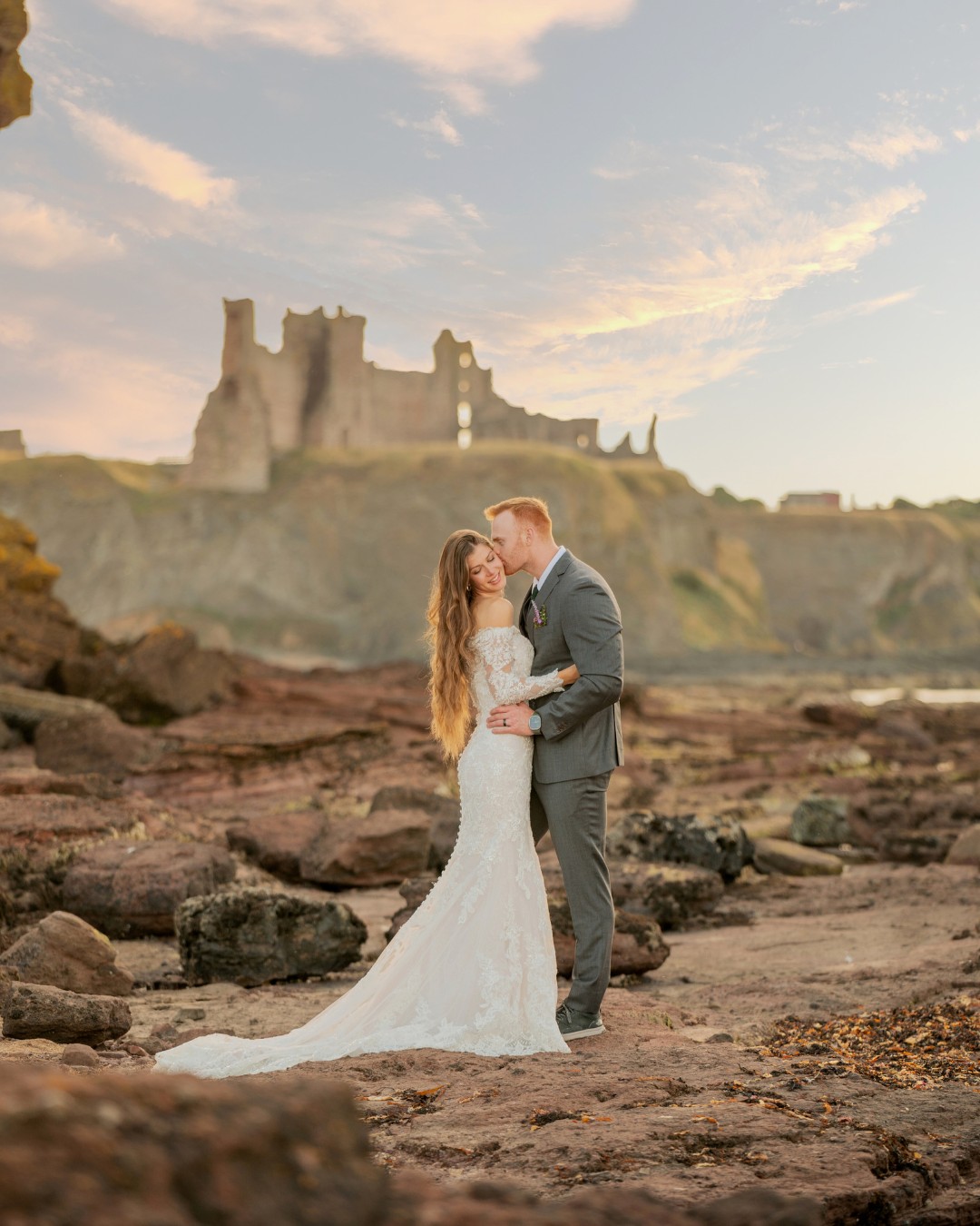 couple stand on Scotland's rocky coast with castle in background