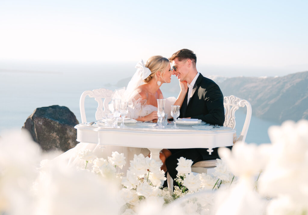 couple sitting at a table with flowers and greek coast backdrop