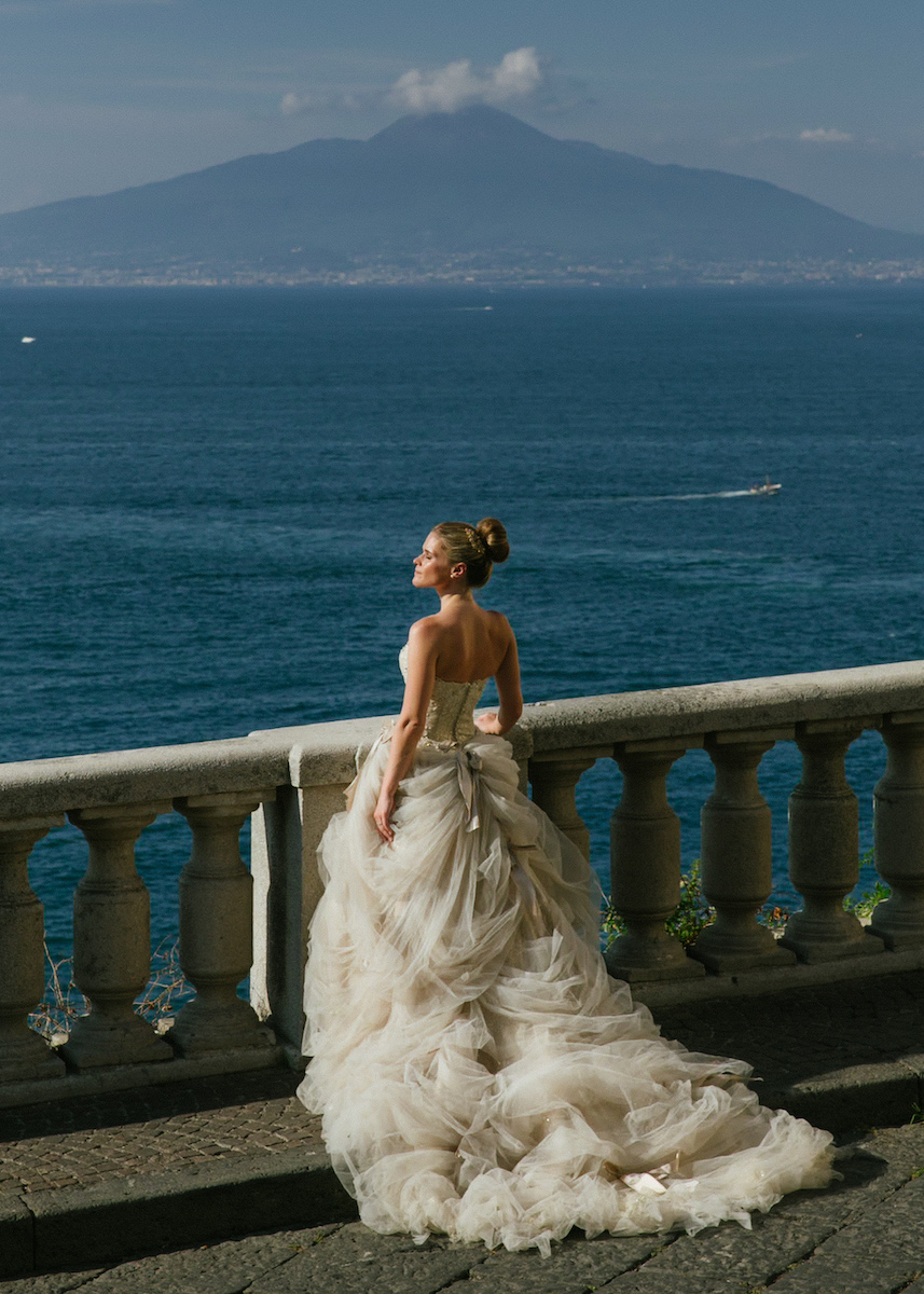 bride poses with bold ball gown dress and backdrop of amalfi coast with mountains behind lake