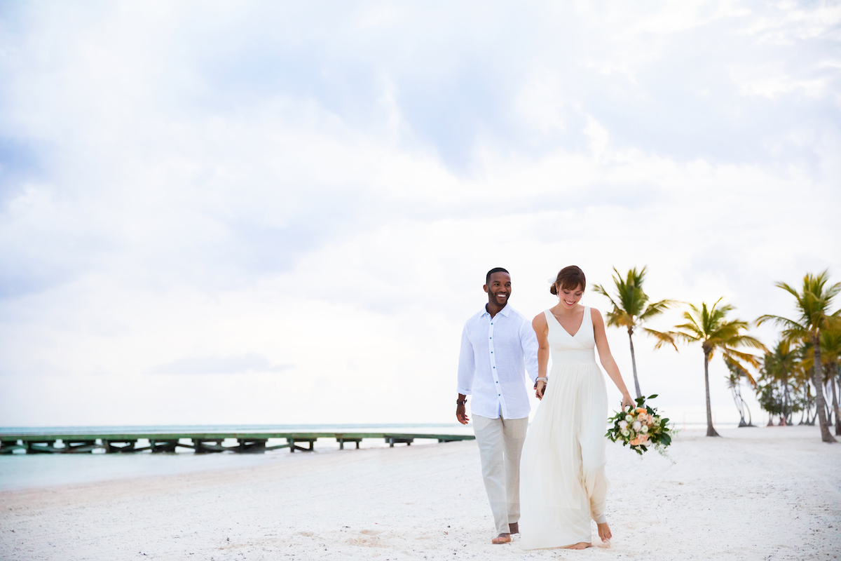 A newlywed couple holding hands and walking on a beach with palm trees and a pier in the background.