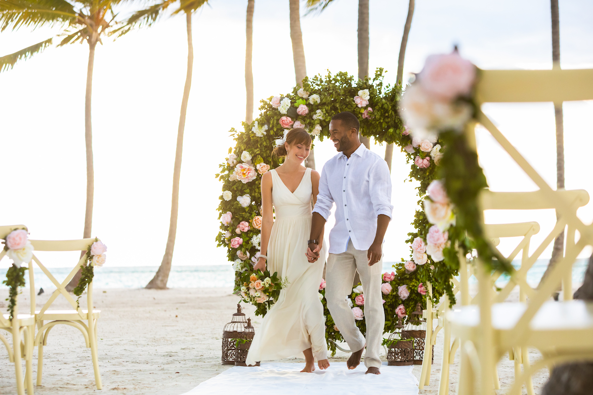 A couple walks hand-in-hand under a floral arch on a sandy beach, with white chairs and palm trees in the background.