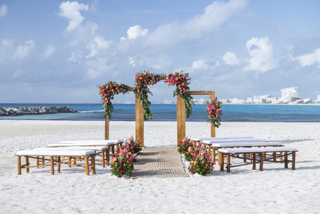 Wooden wedding arch decorated with colorful flowers on a sandy beach with benches, overlooking the ocean and Cancun city skyline in the distance.