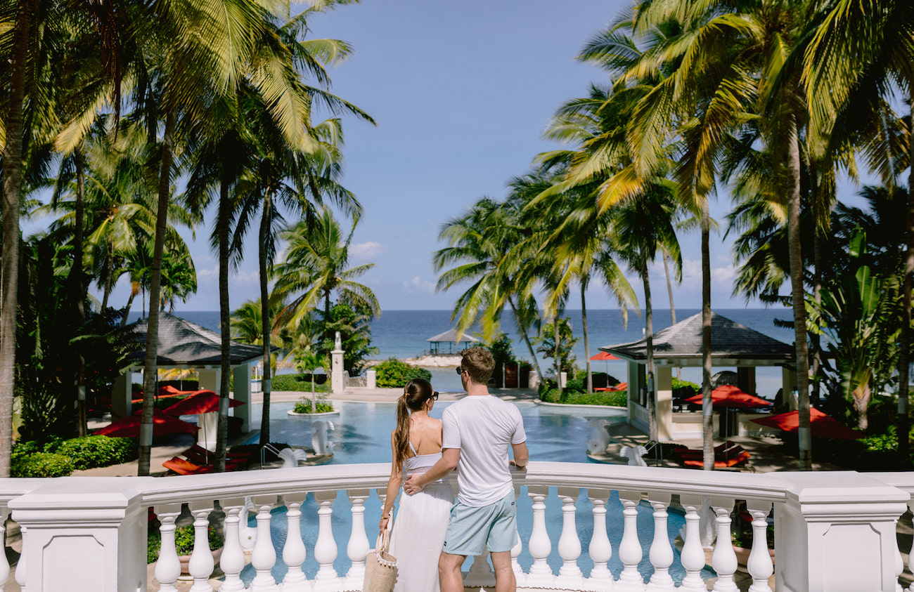 A couple standing together, overlooking Jewel Grande Montego Bay's luxurious pool surrounded by palm trees, facing the ocean.