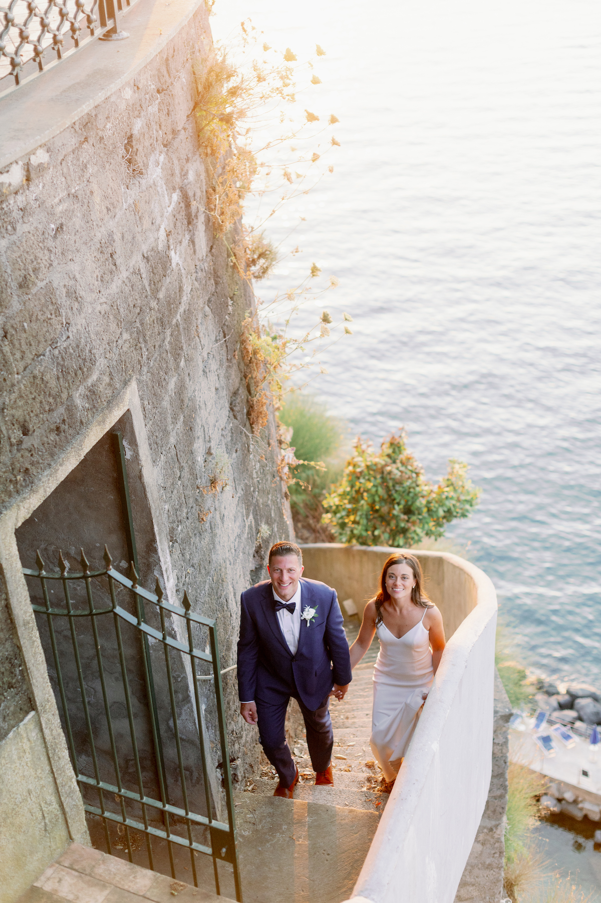 bride and groom beam as they climb side stairs at a lakeside stone building