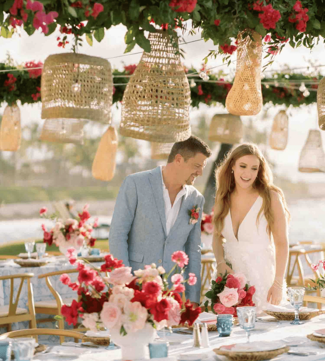 A couple stands at a decorated outdoor table with floral arrangements and hanging wicker lanterns overhead. The man wears a light blue suit, and the woman wears a white dress.