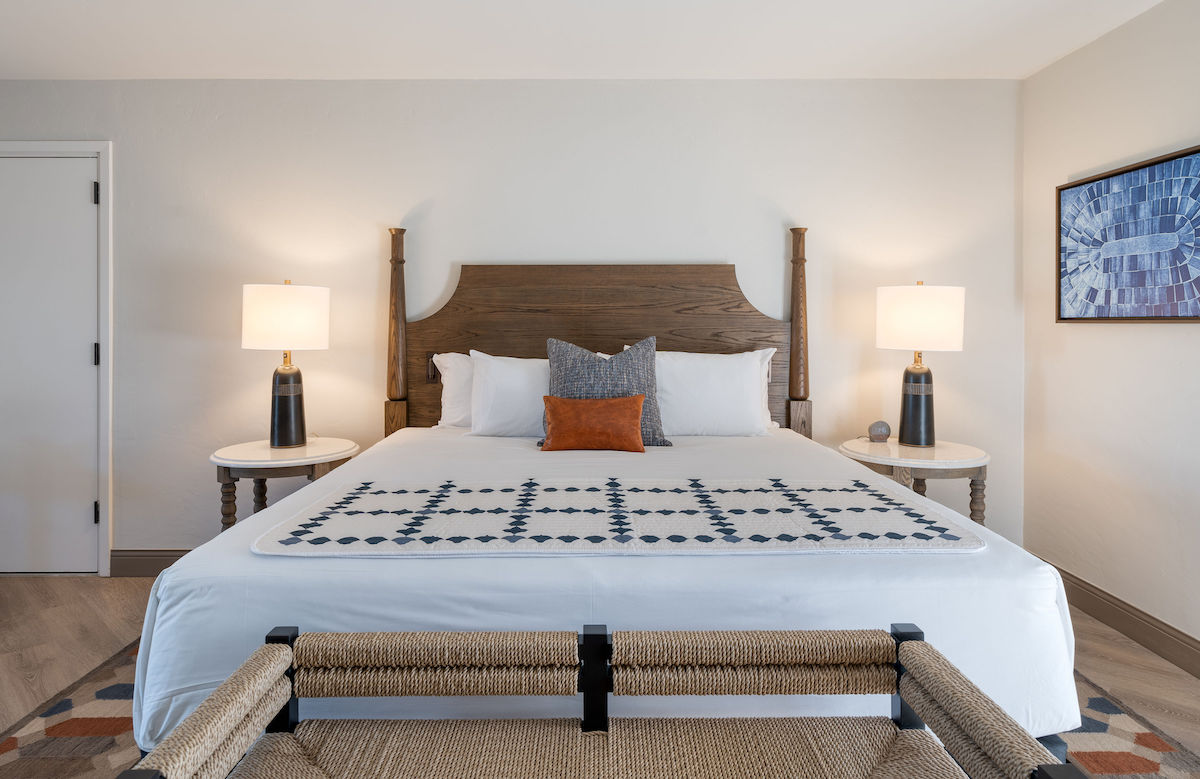 A modern hotel room featuring a king-sized bed with a wooden headboard, two bedside tables with lamps, a bench at the foot of the bed, and a framed blue artwork above the bed.
