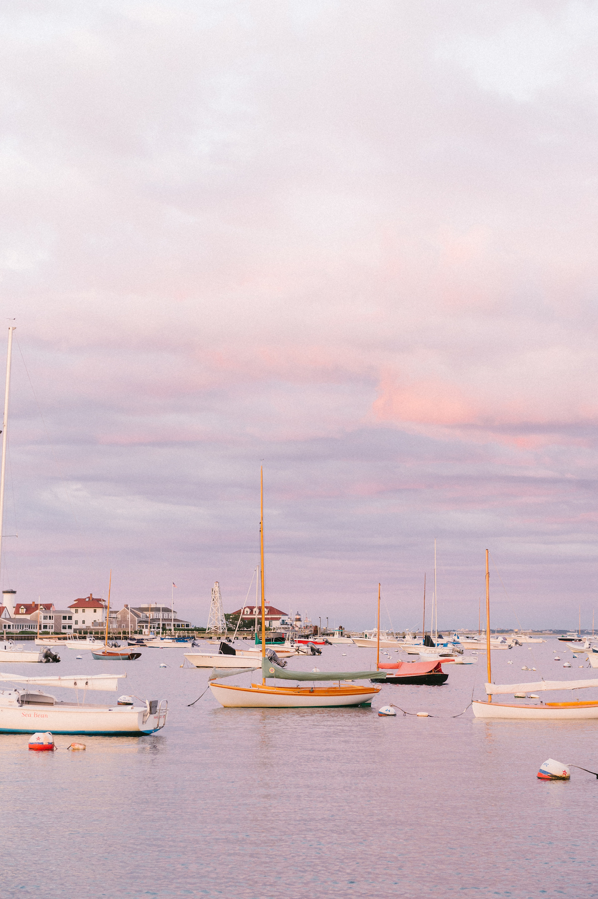 Sailboats moored in a calm harbor at sunset with pastel pink and blue sky, with buildings and a crane in the background.