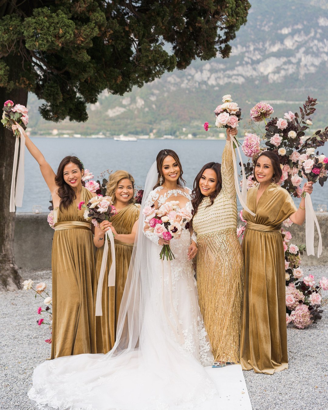 bridesmaids cheer with bride in mustard dresses while holding pink bouquets by Lake Como