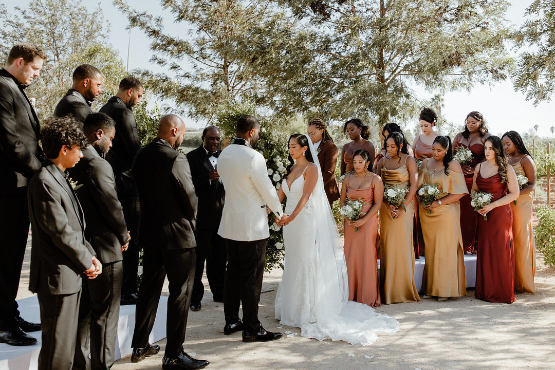 bride and groom stand at altar surrounded by bridesmaids in fall colors and groomsmen in black tuxes at Rancho los Retoños