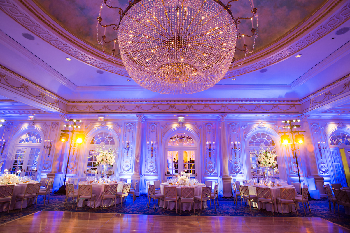 Elegant banquet hall with large crystal chandelier, blue lighting, and tables set for an event. decor includes white floral arrangements.