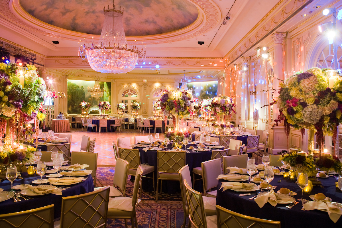 Elegant banquet hall with decorated tables, floral centerpieces, and grand chandeliers, showcasing a luxurious setting for an event.