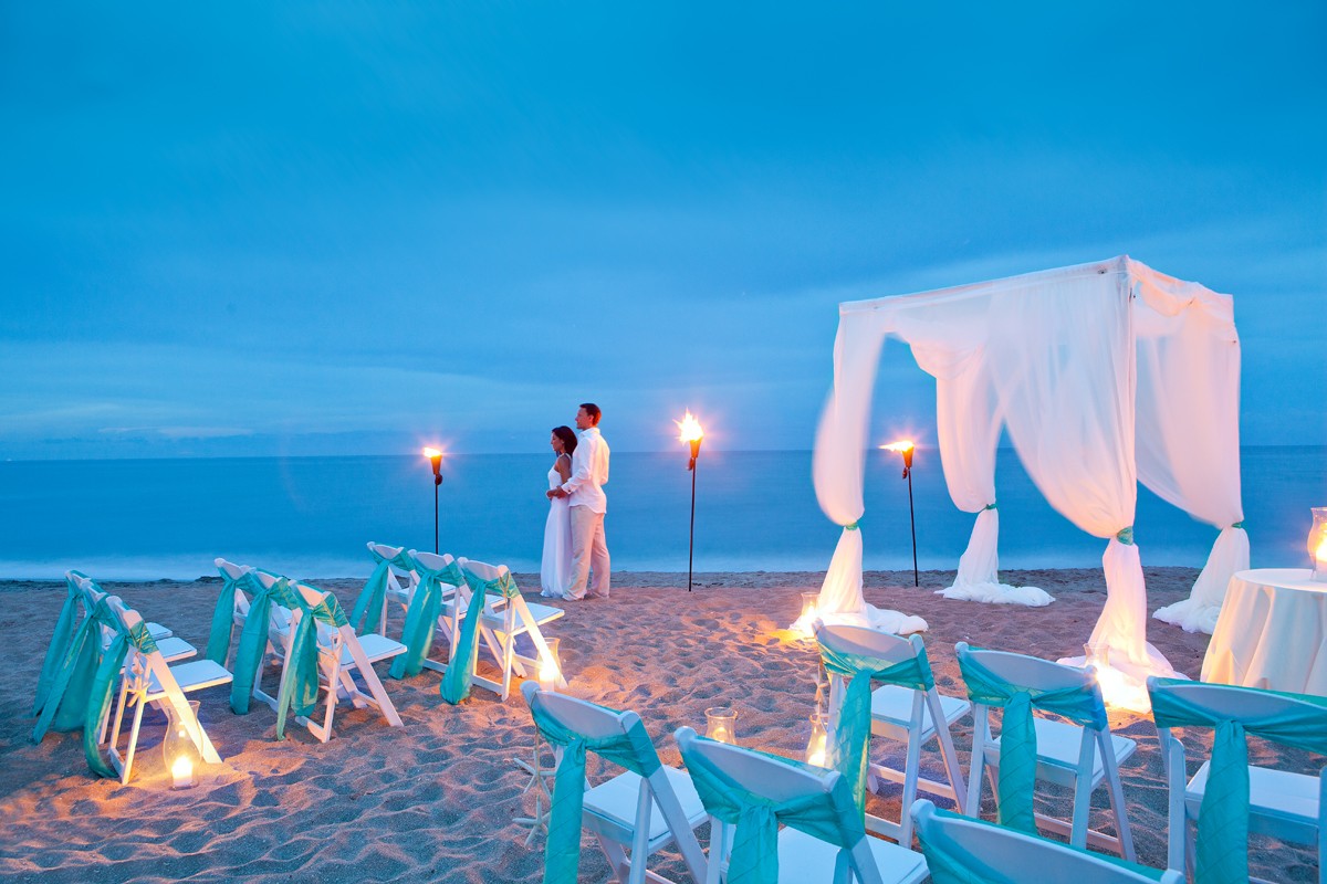 A couple embraces under a white canopy on a sandy beach at dusk, surrounded by white chairs and lit torches, with the ocean in the background.