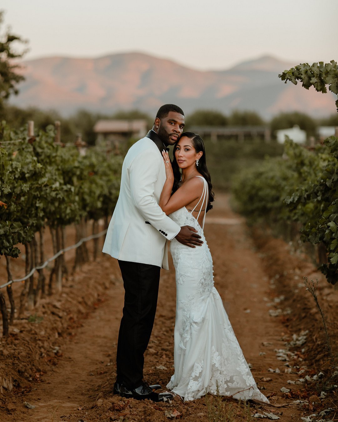 bride and groom hold each other between the vines of Rancho Los Retoños vineyards with the Baja California mountains as a backdrop