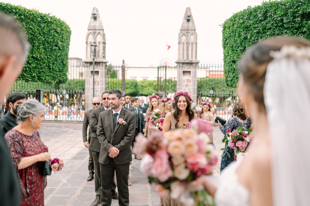 bridesmaids with flower crowns and groomsmen in grey suits look at bride and groom entering San Miguel chapel