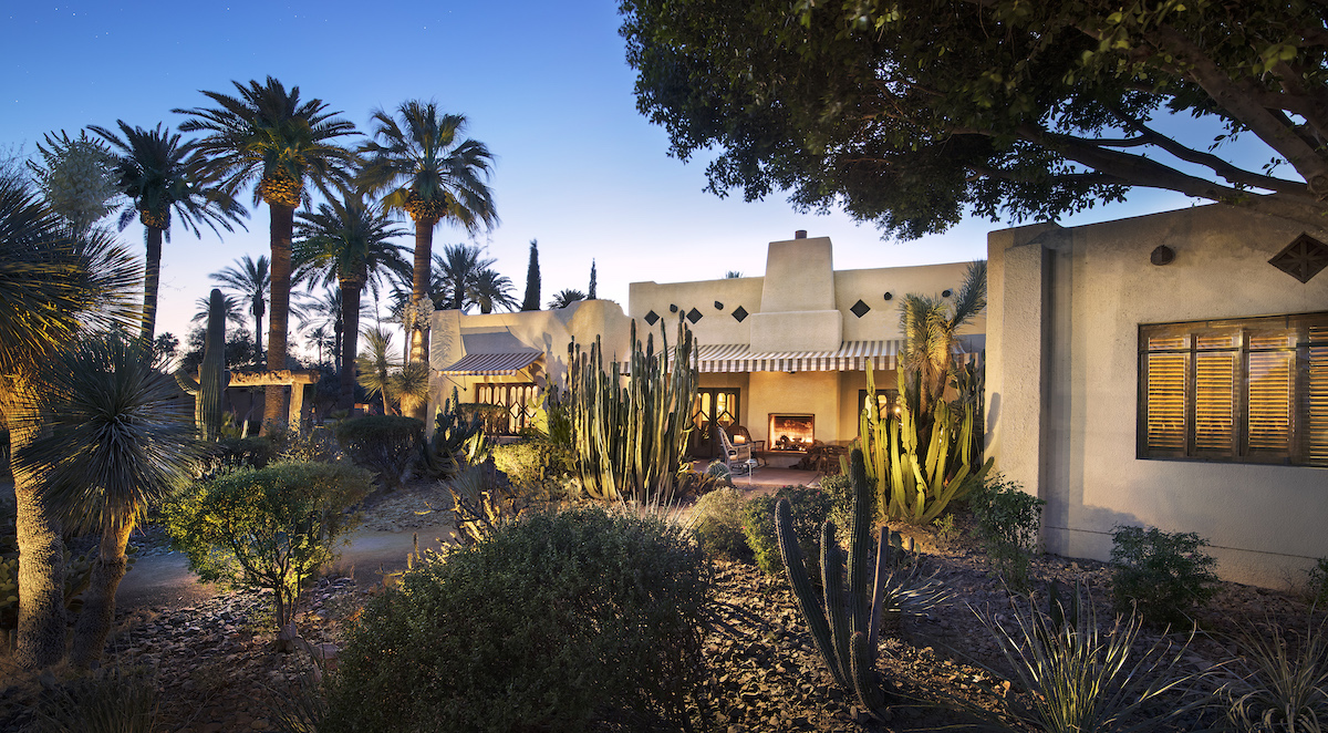 A southwestern-style home surrounded by palm trees and cacti, illuminated in the twilight with visible outdoor lighting.