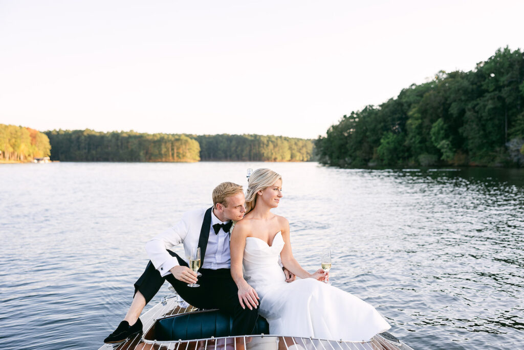 A bride and groom in wedding attire sitting on the back of a boat on a lake, holding champagne glasses and looking into the distance.