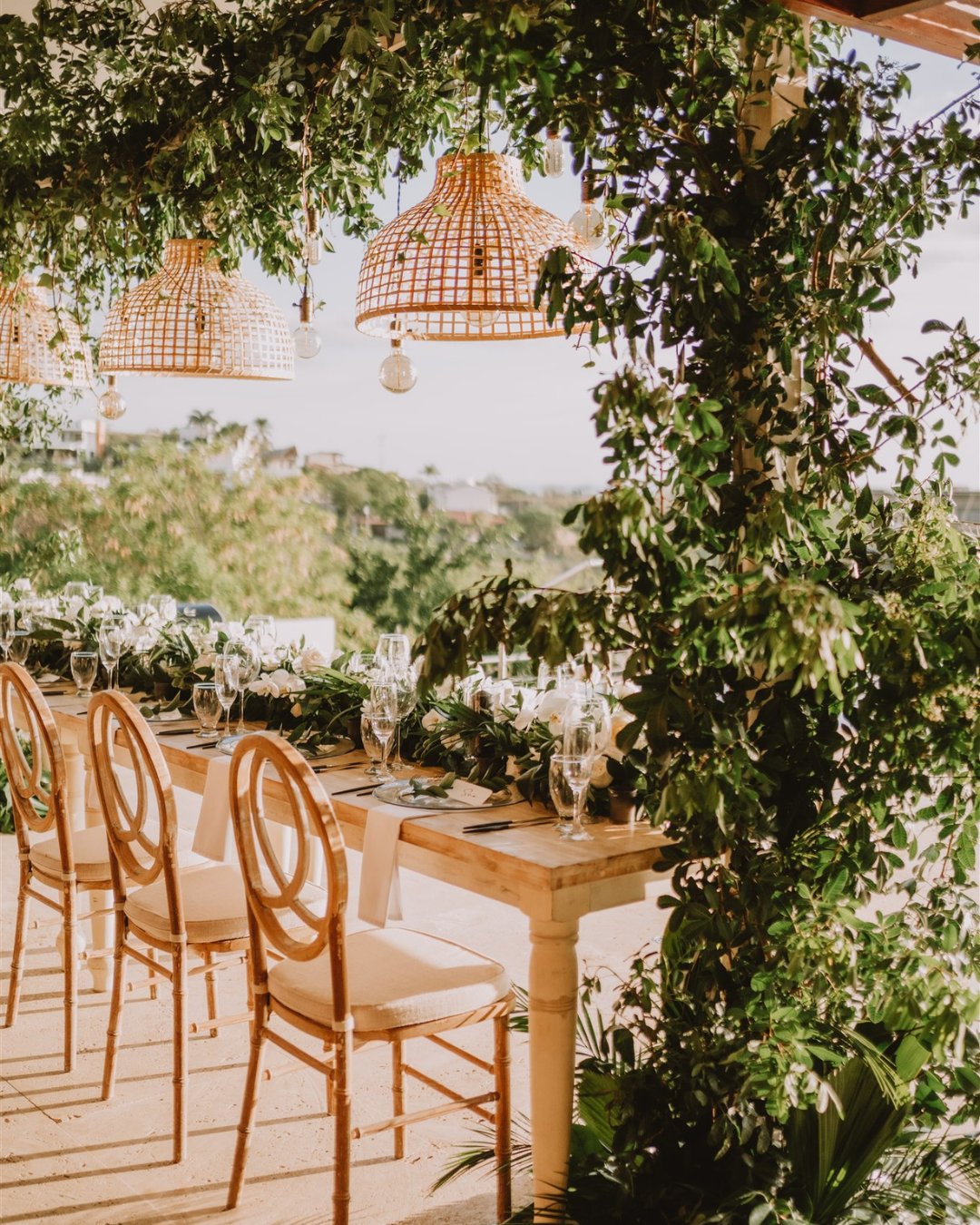 An outdoor dining table set for an event with elegant floral arrangements and wooden chairs overlooks a scenic ocean view at sunset, under hanging wicker lamps and decorative greenery.