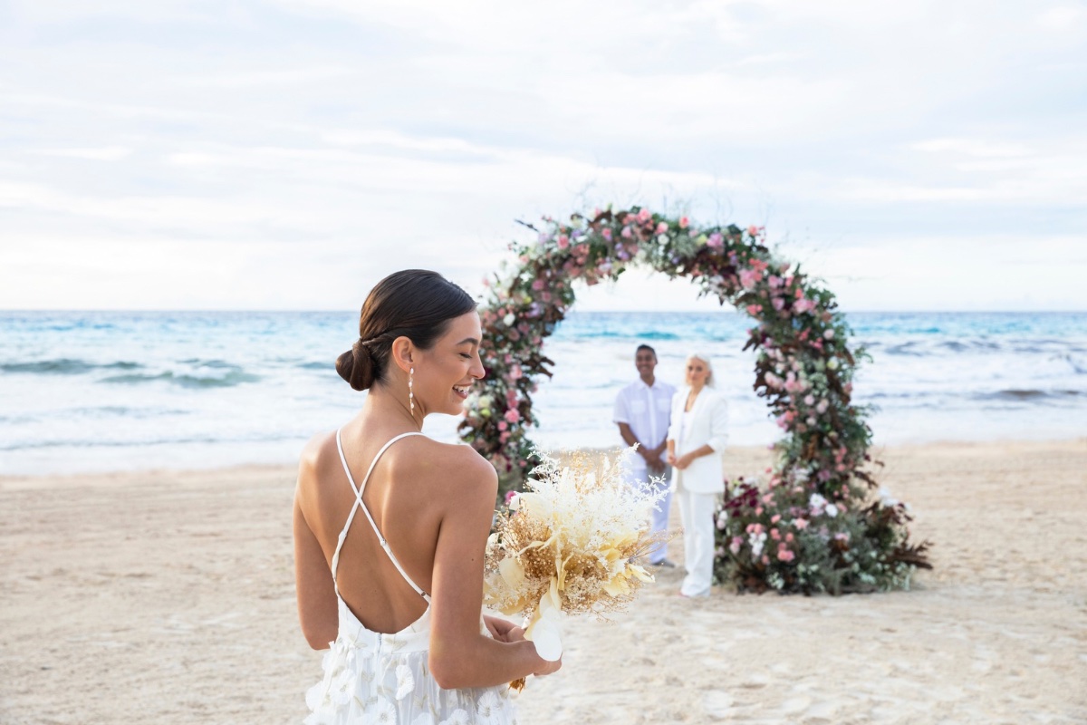 One bride walks down the aisle, towards her bride, who stands under a floral arch with the ocean in the background