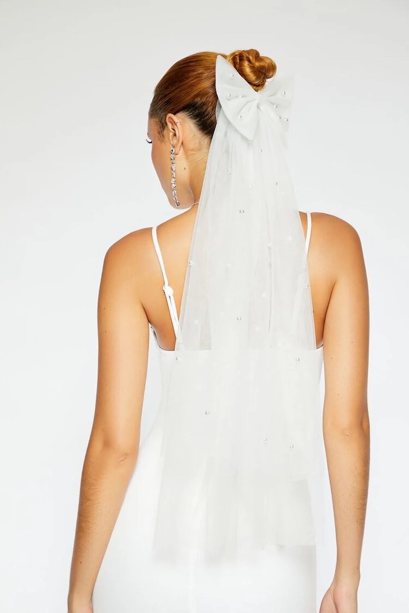 A woman with blond hair in an updo, wearing a Forever 21 white veil adorned with small gems and a large bow, seen from the back. She has earrings and is dressed in a sleeveless white garment.