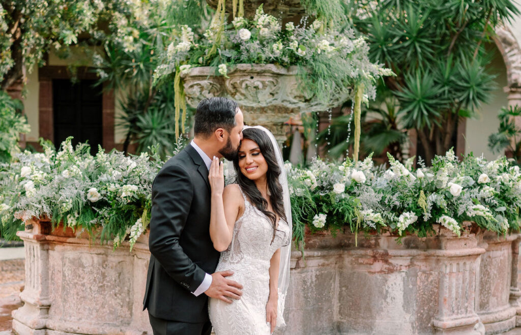 A bride and groom stand in front of a stone fountain adorned with greenery and white flowers. The groom kisses the bride's forehead while she smiles, holding his hand.