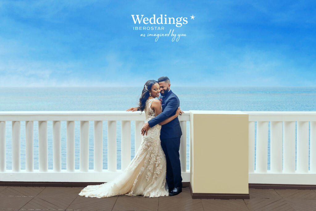 bride and groom hug on terrace overlooking the Caribbean Sea with the Iberostar Weddings logo