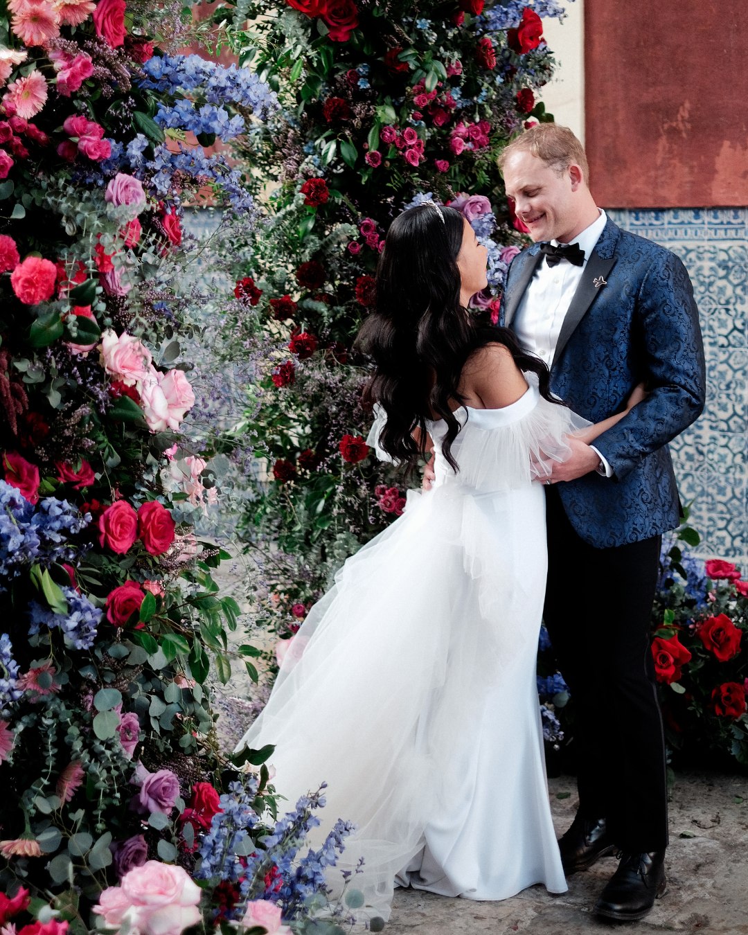 Roraine and Tyler look at each other under a pink and blue floral arch
