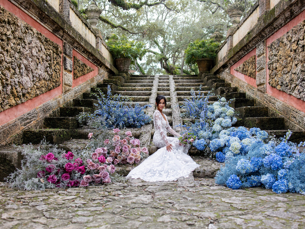 bride sits at bottom of stairs surrounded by cloud-like blue and pink floral arrangements