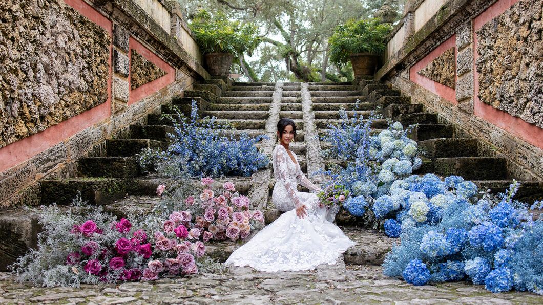 bride sits at bottom of stairs surrounded by cloud-like blue and pink floral arrangements