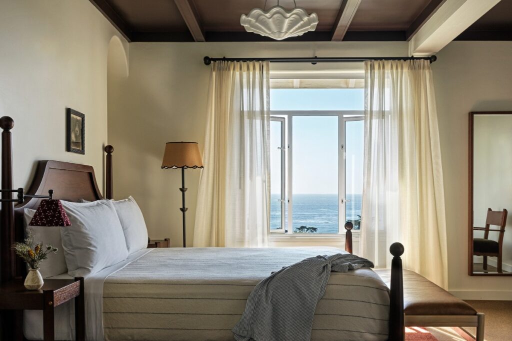 A bedroom with a king-size bed facing a large window overlooking the ocean. The bed has a white duvet and blue pillows. There is a nightstand on either side of the bed with lamps on top. In the corner of the room is a armchair. The room has white walls and a cream colored rug.