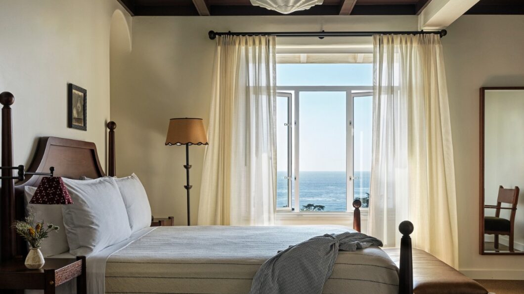 A bedroom with a king-size bed facing a large window overlooking the ocean. The bed has a white duvet and blue pillows. There is a nightstand on either side of the bed with lamps on top. In the corner of the room is a armchair. The room has white walls and a cream colored rug.