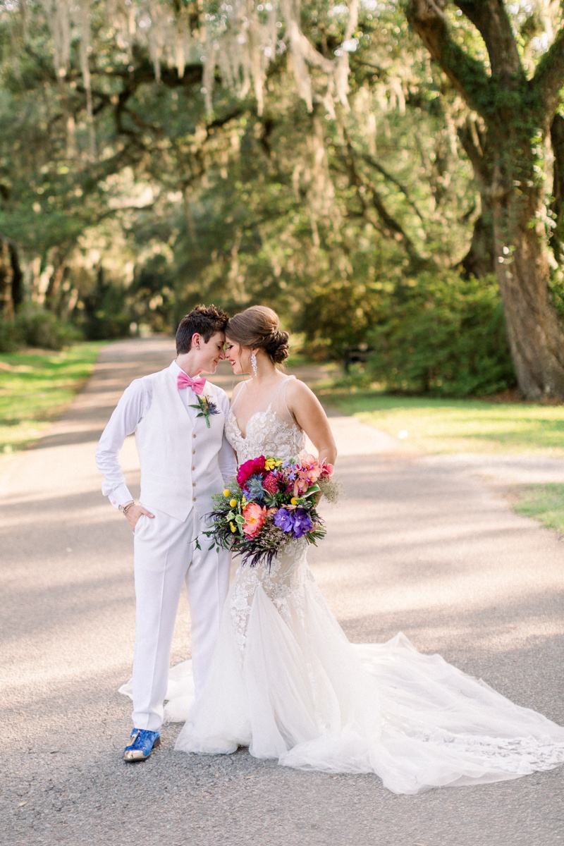 Two people in wedding attire stand closely and touch foreheads on a tree-lined road, one wearing a white suit and the other a white gown holding a colorful bouquet.