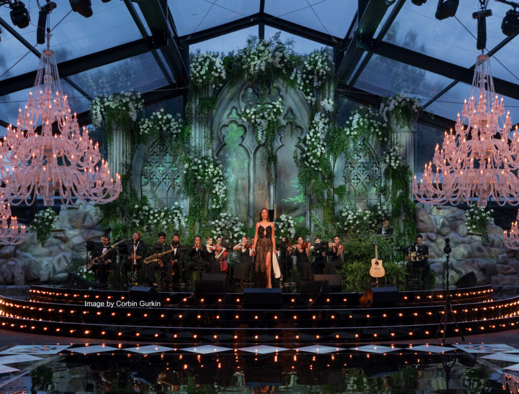 A concert stage is adorned with lush greenery, floral decorations, and elegant chandeliers. A female performer stands center stage surrounded by musicians playing string instruments, with arched, gothic-style windows in the backdrop, creating a serene ambiance.