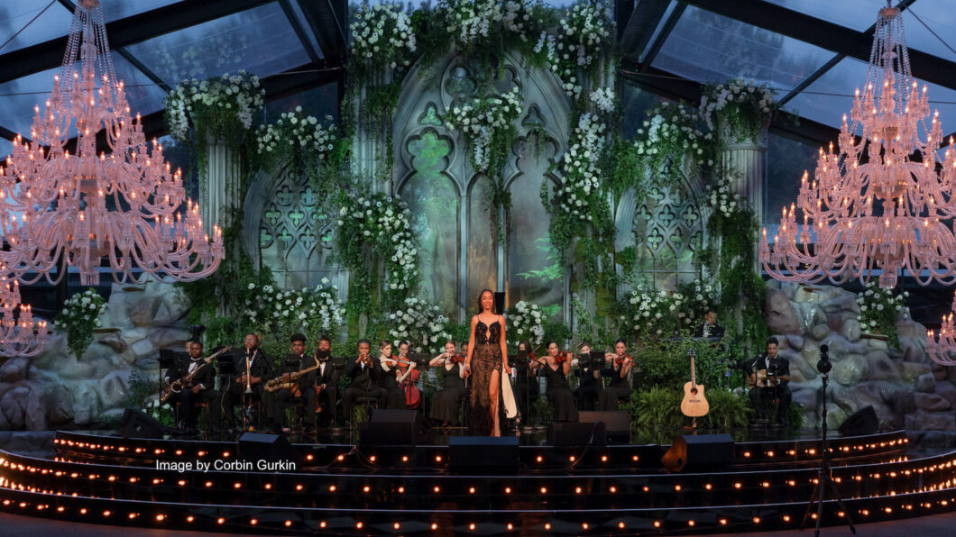 A concert stage is adorned with lush greenery, floral decorations, and elegant chandeliers. A female performer stands center stage surrounded by musicians playing string instruments, with arched, gothic-style windows in the backdrop, creating a serene ambiance.