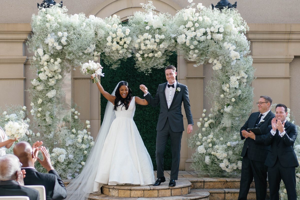 Geornay and Tyler celebrate joyously under a floral archway after their wedding ceremony. The bride wears a white gown and veil, holding a bouquet, while the groom wears a suit. Guests and officiants applaud them, with an elegant background setting.
