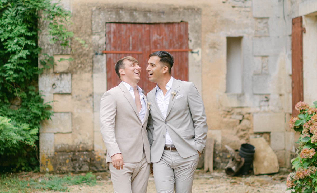 Noel and Daniel laugh together walking shoulder to shoulder away from an old French countryside building with a red door