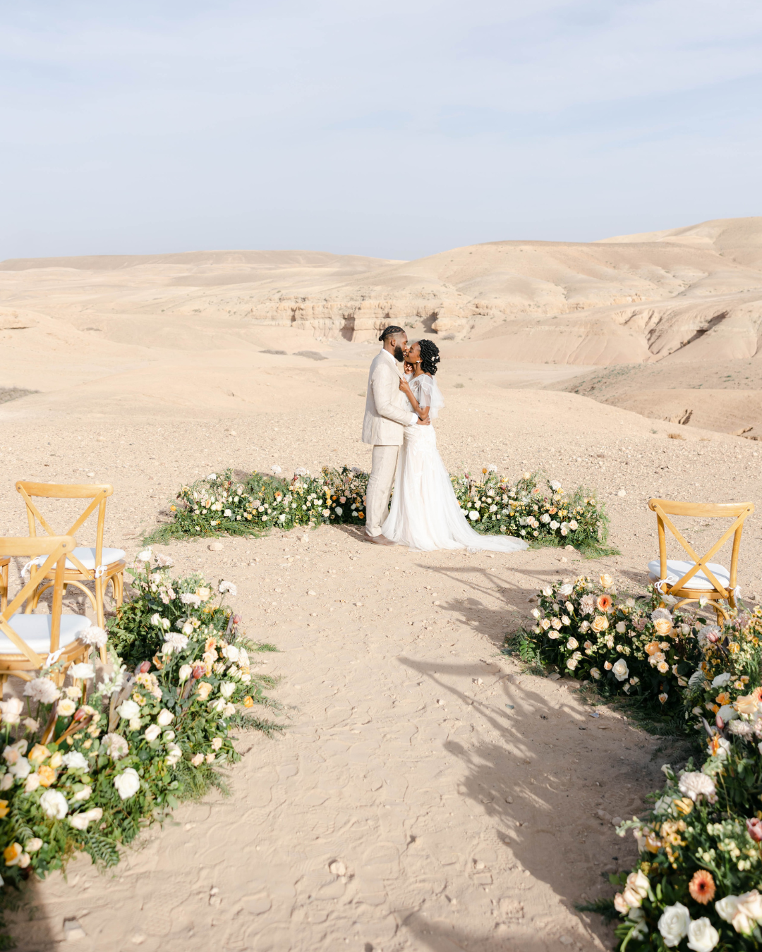 A bride and groom stand facing each other and holding hands in the center of a floral arrangement on a desert landscape. Two wooden chairs with white cushions are positioned on either side of the path leading to the couple. The sky is clear and blue.