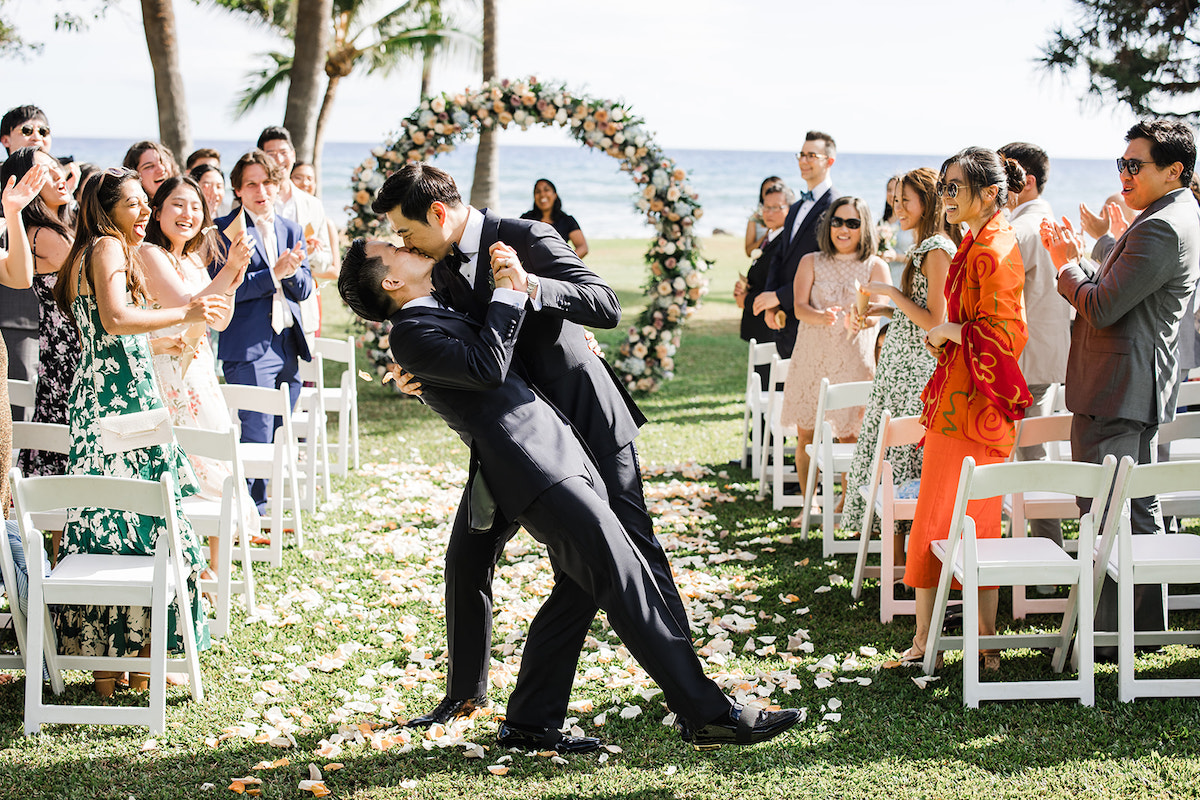 groom dips and kisses his groom with wedding guests clapping to the side and circle floral arch and ocean in background