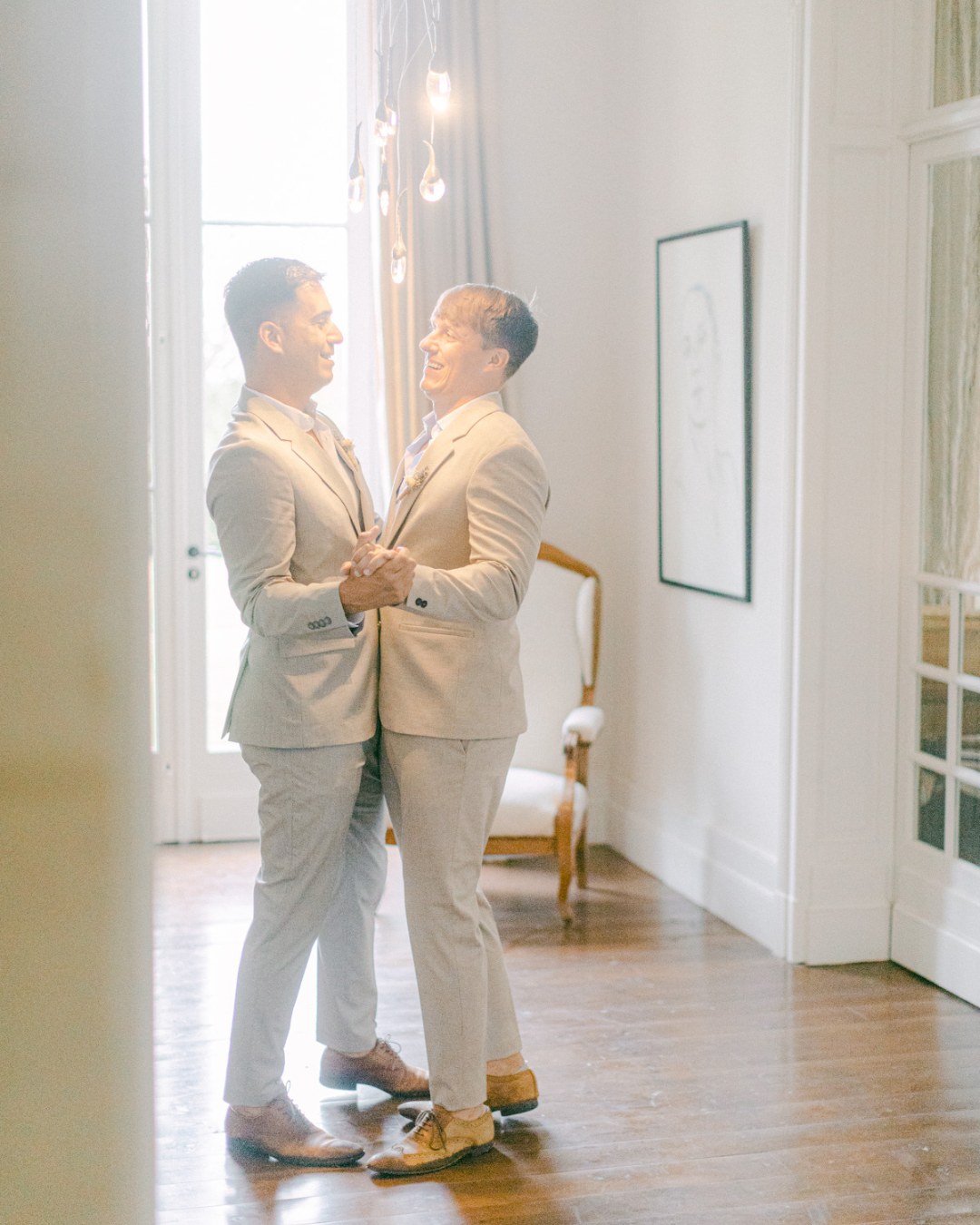 Noel and Daniel joyfully dance together in a bright, elegant room with wooden flooring. A chair and a framed sketch are visible in the background, and the room is softly lit by hanging light bulbs.