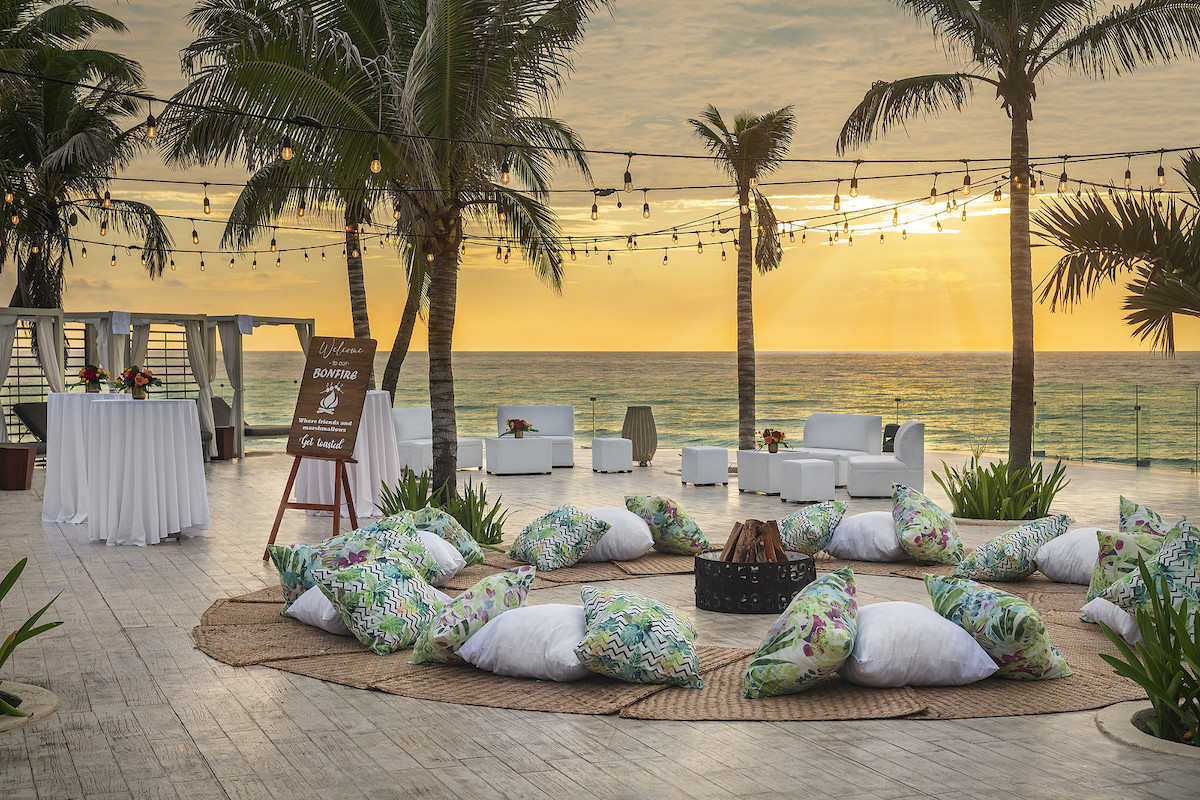 A bonfire on a beach at sunset. The bonfire is surrounded by colorful pillows for seating.