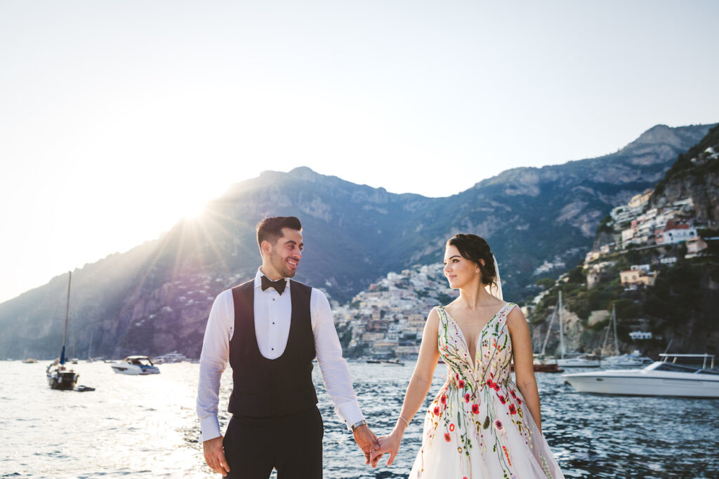 A couple stands hand in hand by the water, dressed in wedding attire. The groom wears a black tuxedo, and the bride wears a floral gown. They look at each other lovingly with a picturesque coastal town and mountains in the background. Boats float nearby.