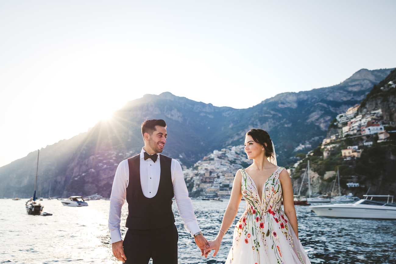 A couple stands hand in hand by the water, dressed in wedding attire. The groom wears a black tuxedo, and the bride wears a floral gown. They look at each other lovingly with a picturesque coastal town and mountains in the background. Boats float nearby.