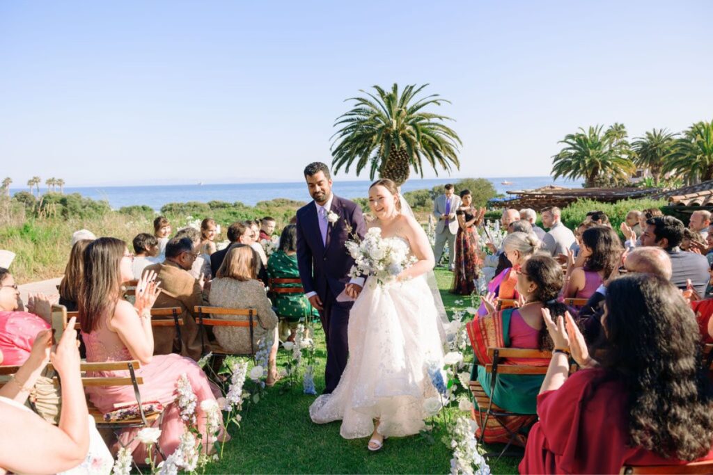 A newlywed couple walks down an outdoor aisle, surrounded by seated guests. The bride is wearing a white wedding dress and holding a bouquet, while the groom is in a dark suit. The ocean and palm trees can be seen in the background under a clear blue sky.