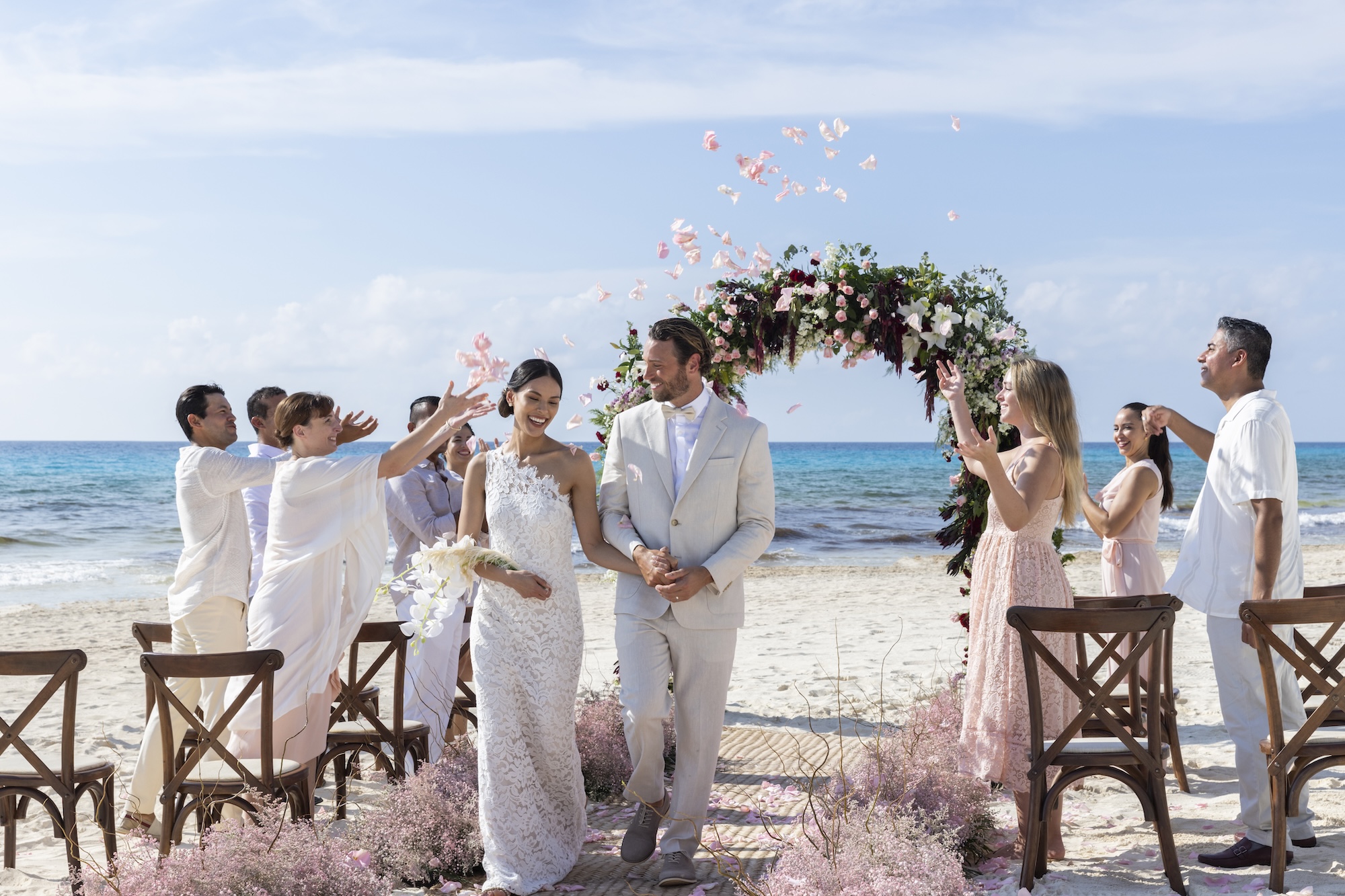 A newlywed couple walks down the aisle on a beach, surrounded by guests throwing petals. The ocean is visible in the background.