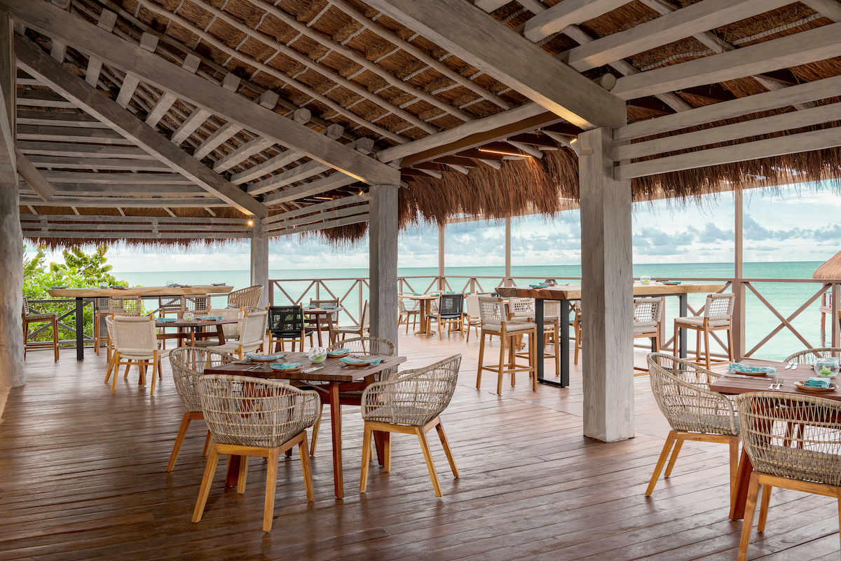 An open-air restaurant with wooden flooring and thatched roof, featuring wooden tables and wicker chairs. The dining area overlooks a scenic ocean view with a cloudy sky. The space is airy and well-lit, creating a relaxed and inviting atmosphere.