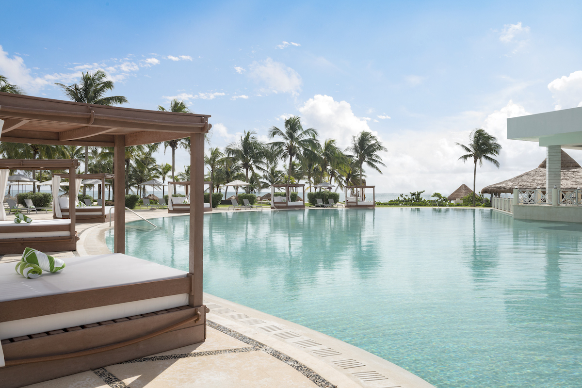 A luxurious resort pool area featuring wooden canopy beds with white cushions. The expansive pool is surrounded by palm trees and cabanas under a bright, clear sky, creating a serene and inviting tropical atmosphere. In the background, the ocean is visible.